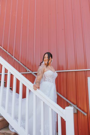 Smiling bride in strapless lace wedding gown with tattoos descends white staircase against red barn wall.