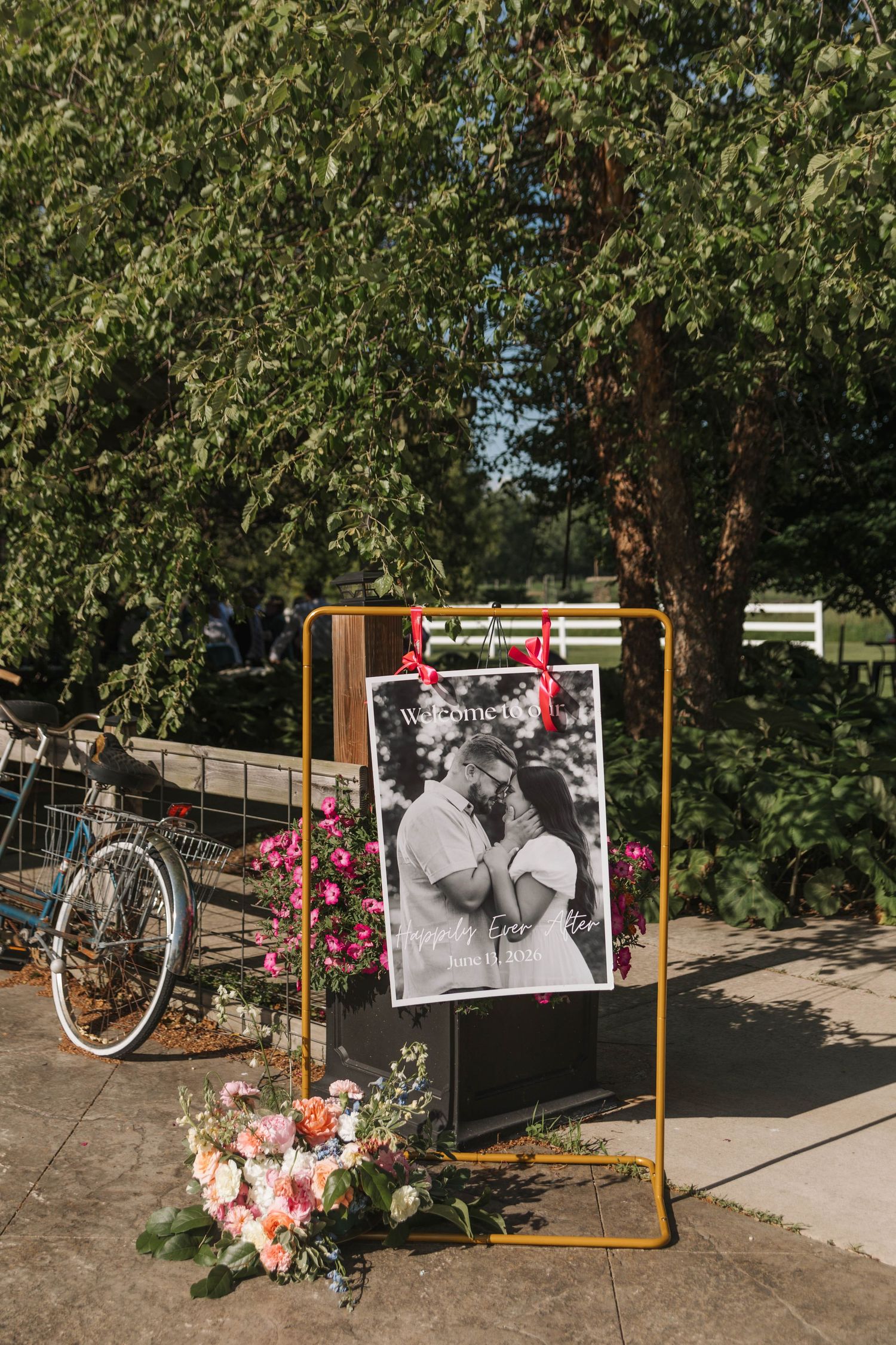 Gold rack wedding welcome sign with couple photo, pink florals, and bicycle in lush garden setting.