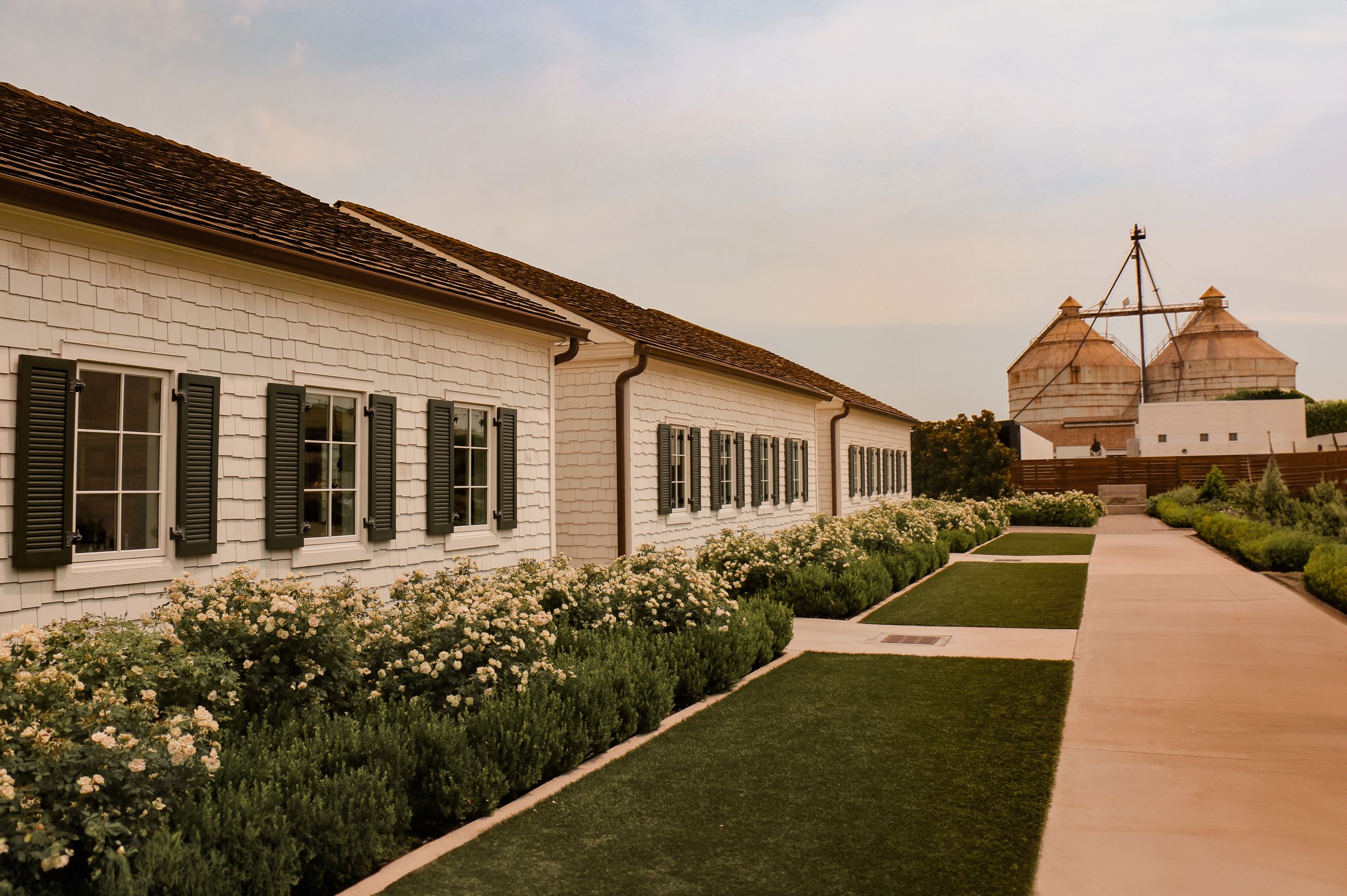 White colonial style building with black shutters and manicured garden pathway leading to a barn structure in distance.