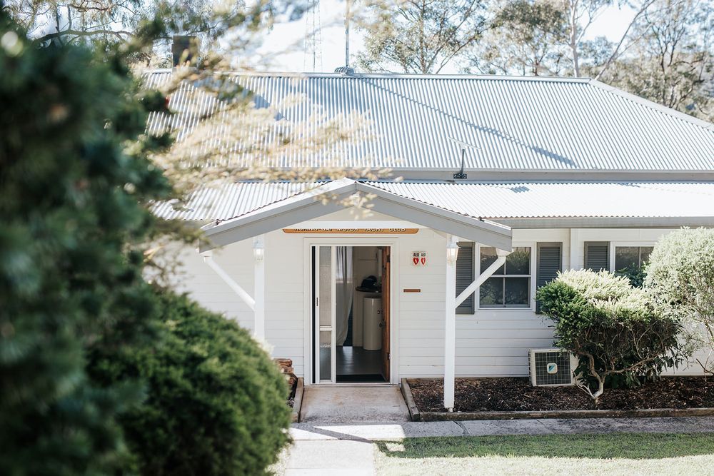 White cottage-style home with grey metal roof, white columns, open front door, and lush green hedges in a serene garden setting.