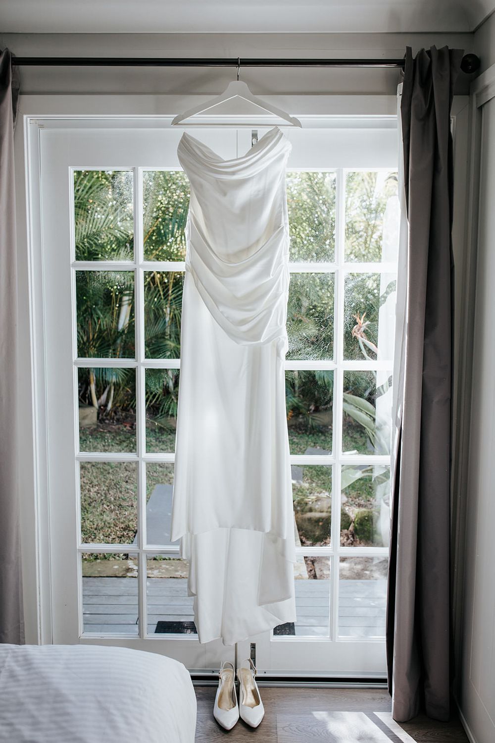 Elegant white satin strapless wedding dress hanging on a window with white heels below.