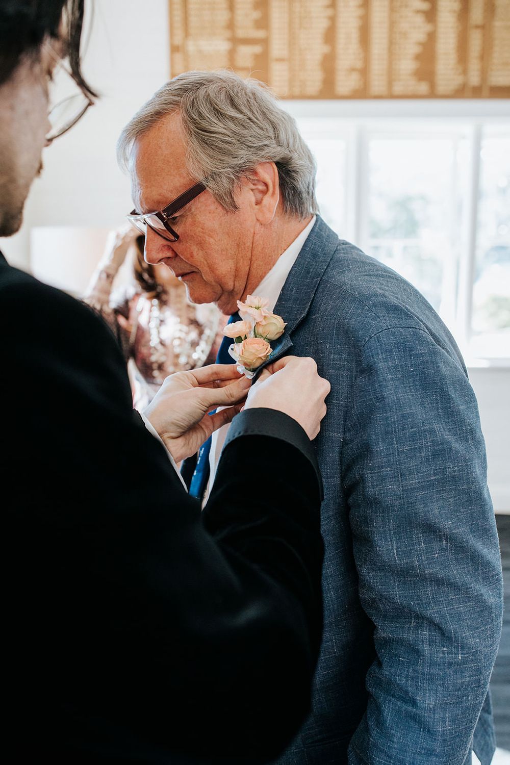 Groomsman pinning a delicate peach rose boutonniere onto an older gentleman in a blue linen suit at a wedding.