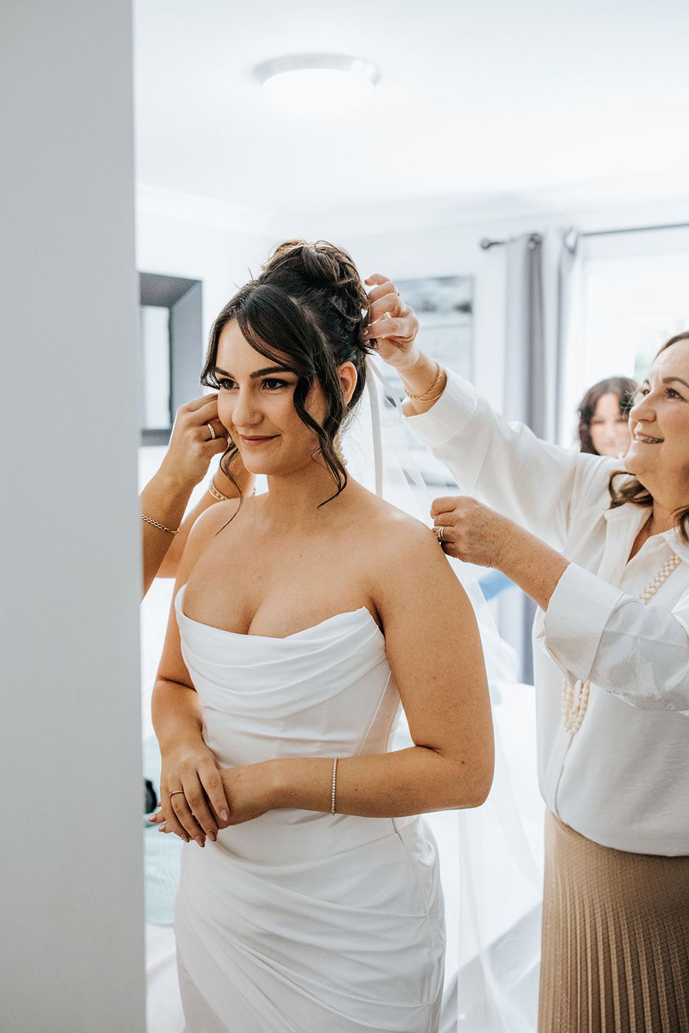 Bride in strapless white gown gets ready as helpers fasten her dress and adjust her earrings indoors.