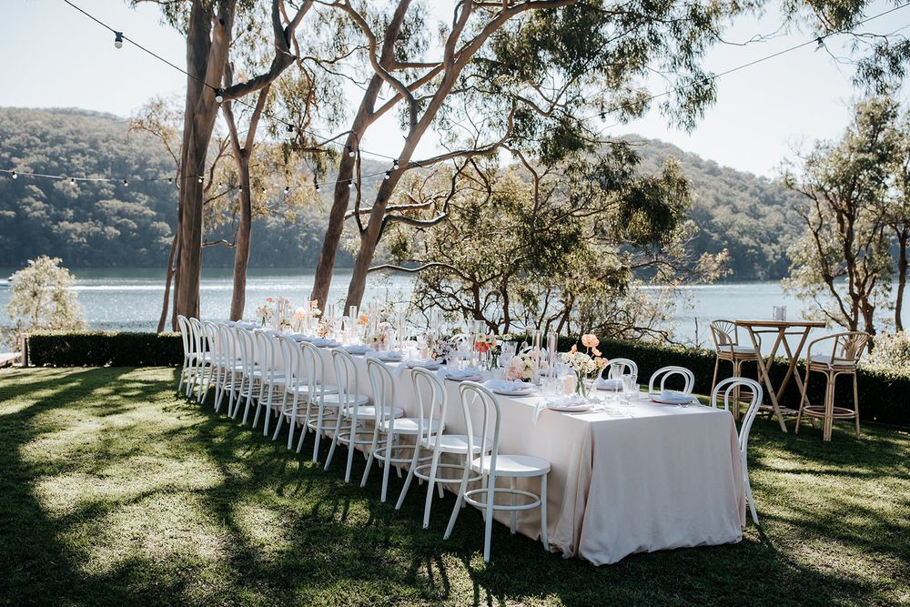 Outdoor waterfront wedding reception with long white table set among trees overlooking a lake.