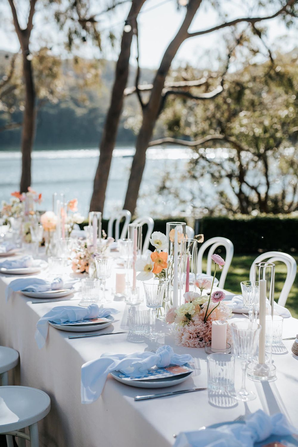 Elegant outdoor wedding reception table with white linens, pastel florals, candles, and a waterfront backdrop.