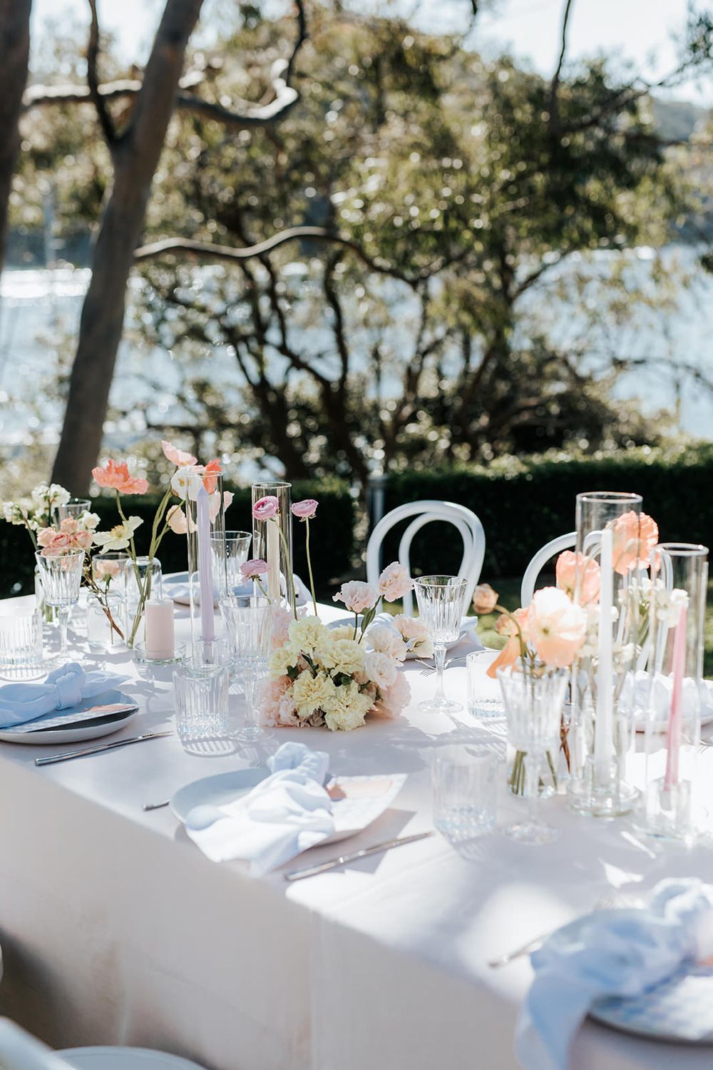 Elegant outdoor wedding table setting with pink flowers, glass vases, and soft blue napkins overlooking a waterfront.