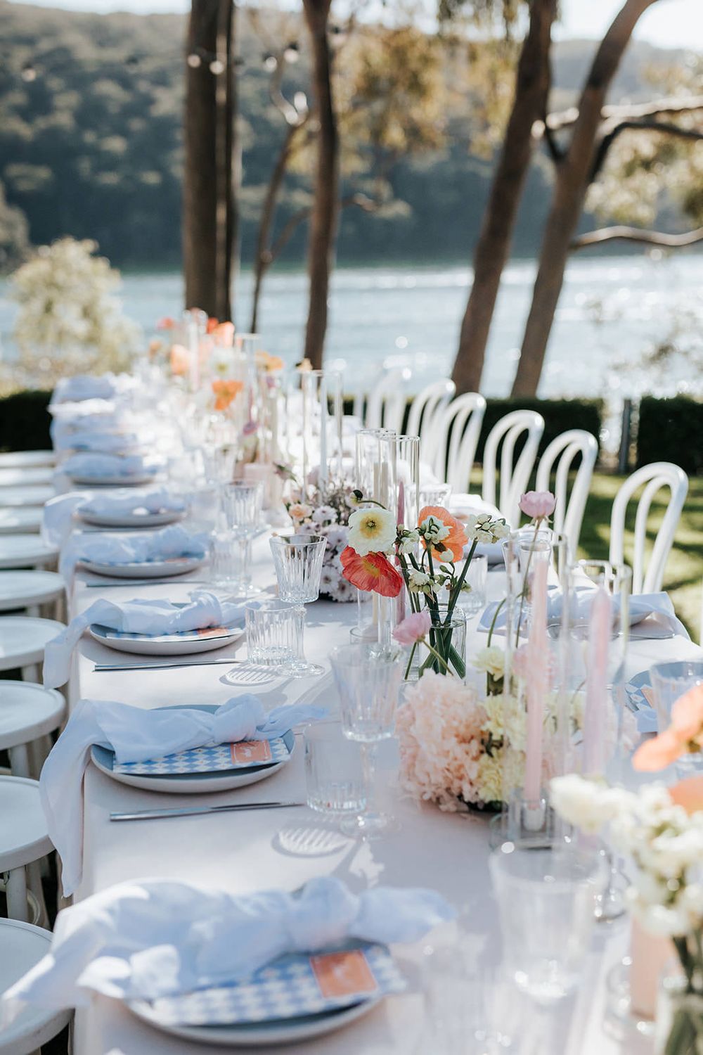 Elegant outdoor wedding reception table set with floral centerpieces, crystal glassware, and blue napkins overlooking a waterfront.