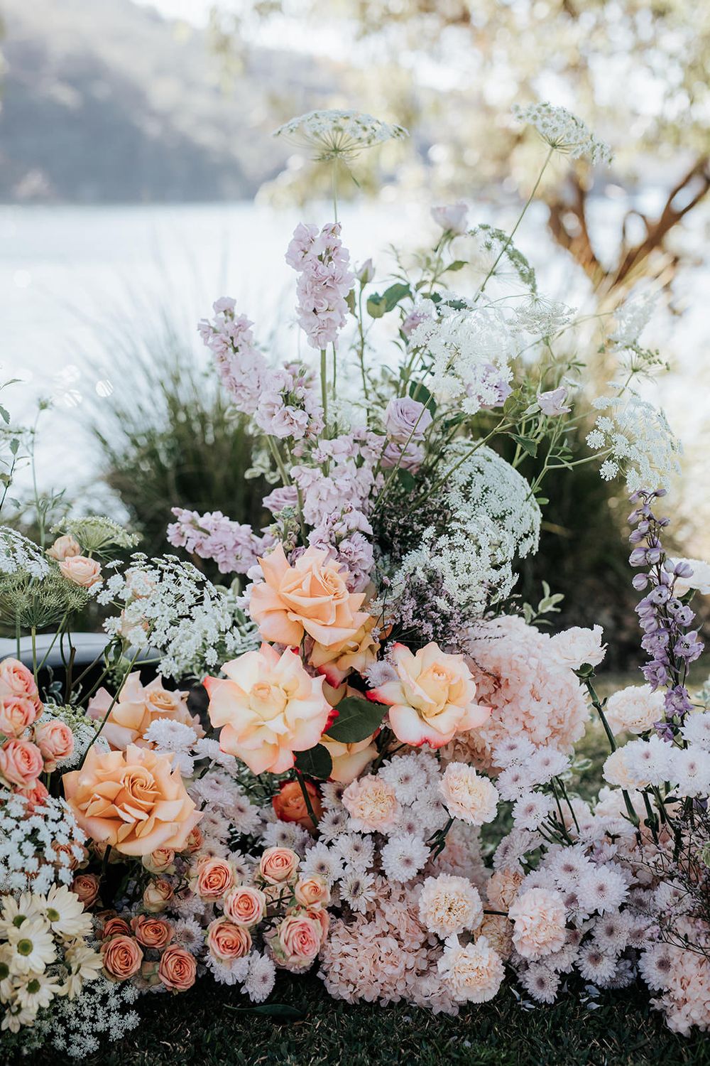 Lush wedding floral arrangement featuring peach roses, lavender blooms, and white wildflowers set against a waterfront backdrop.
