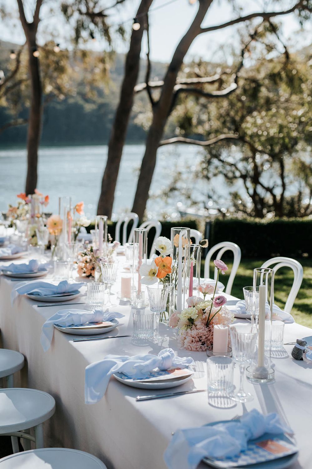 Elegant outdoor wedding reception table set with floral arrangements, candles, blue napkins, and glassware overlooking a serene waterway.