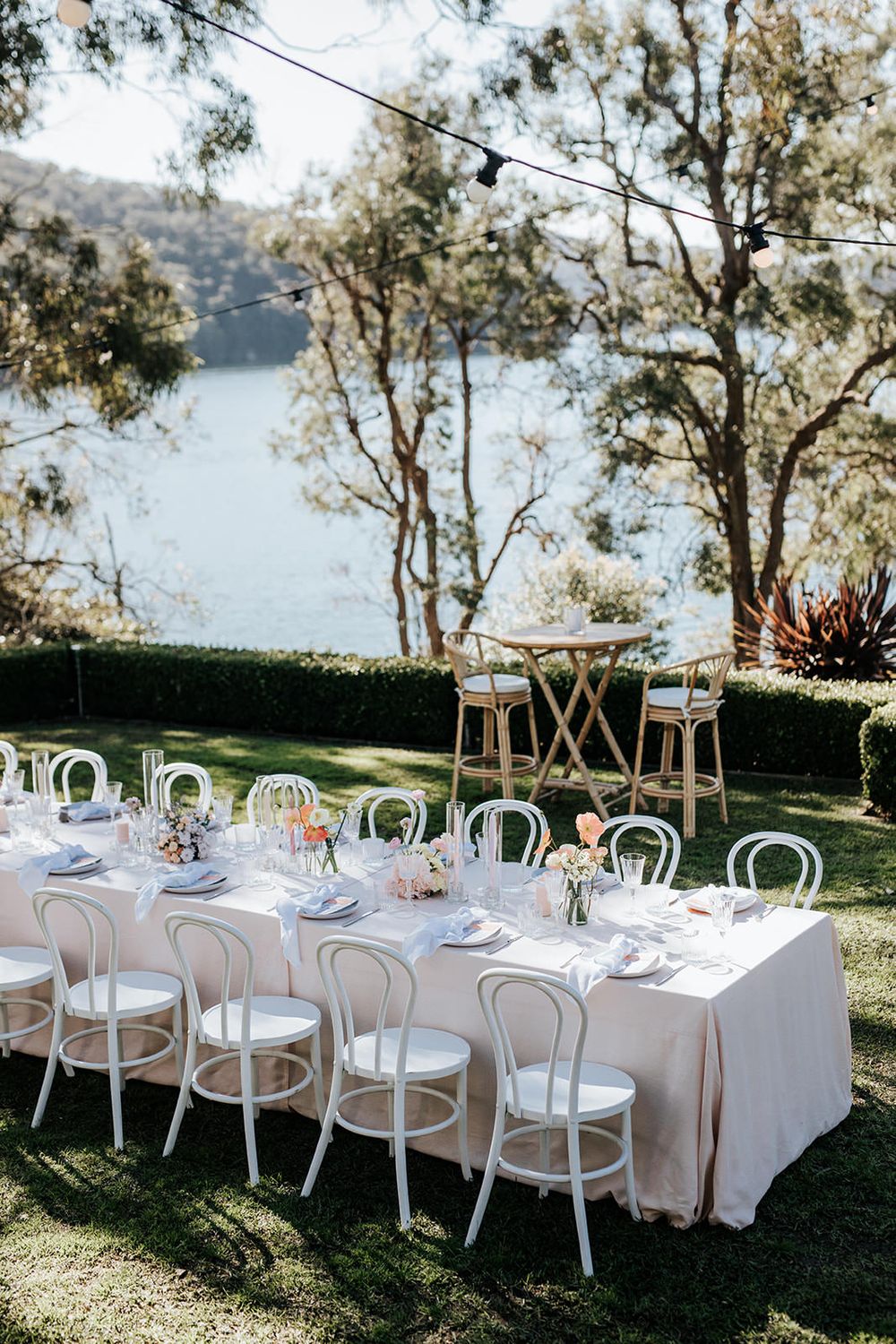 Elegant outdoor wedding reception table set with white bentwood chairs and blush linens overlooking a serene lake.