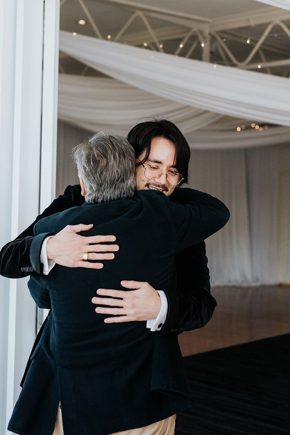 Emotional groom hugging an older man at a wedding reception under draped white fabric ceiling.
