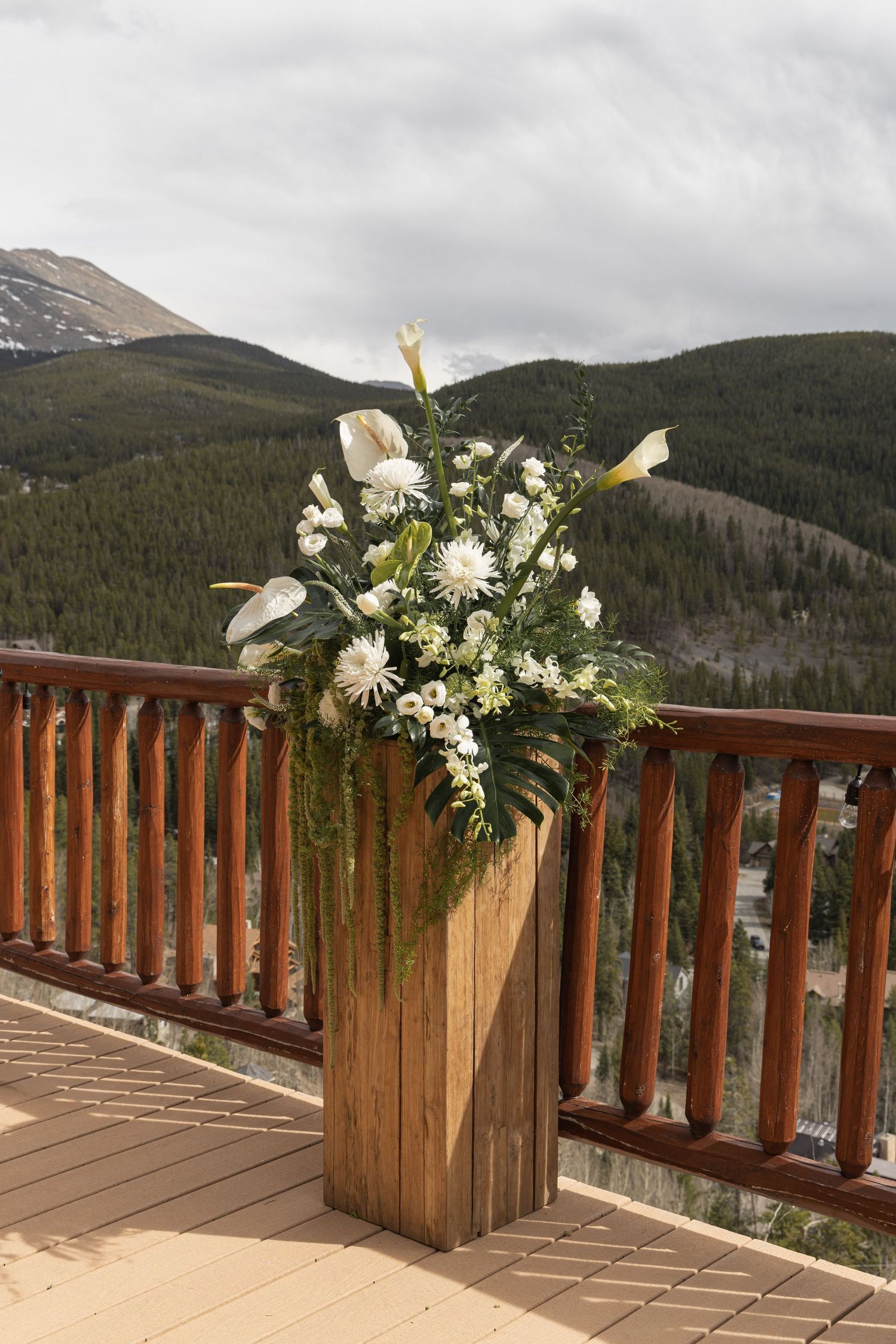 White floral arrangement on wooden pedestal on mountain deck with pine forest backdrop.