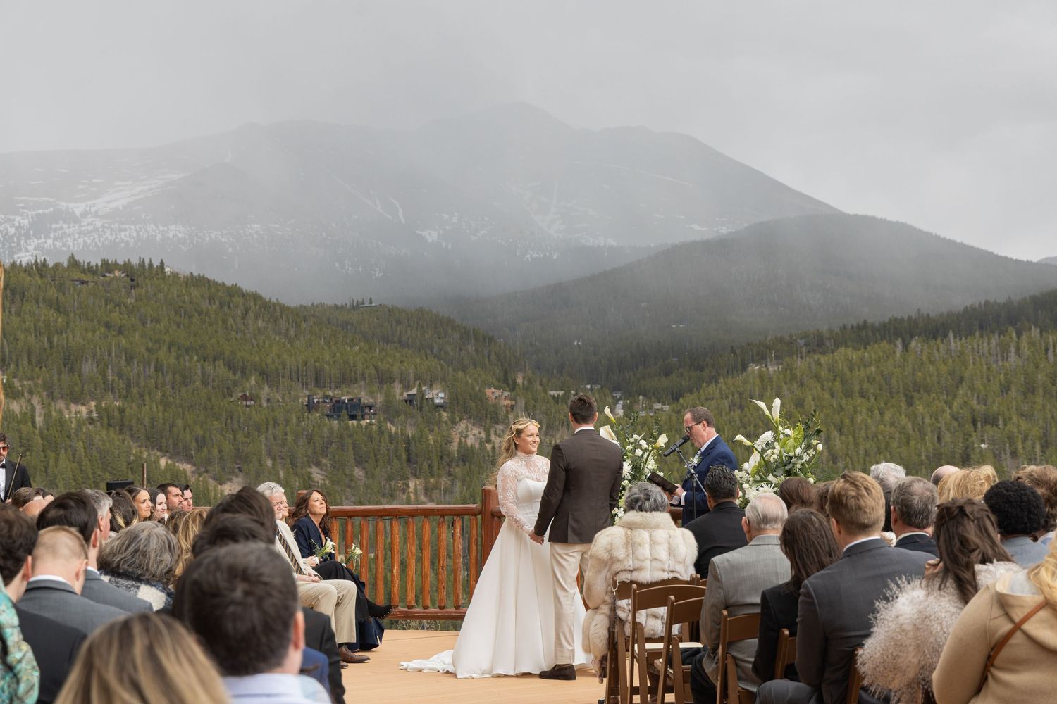 Outdoor mountain wedding ceremony on a deck with pine forests and snow-capped peaks backdrop.