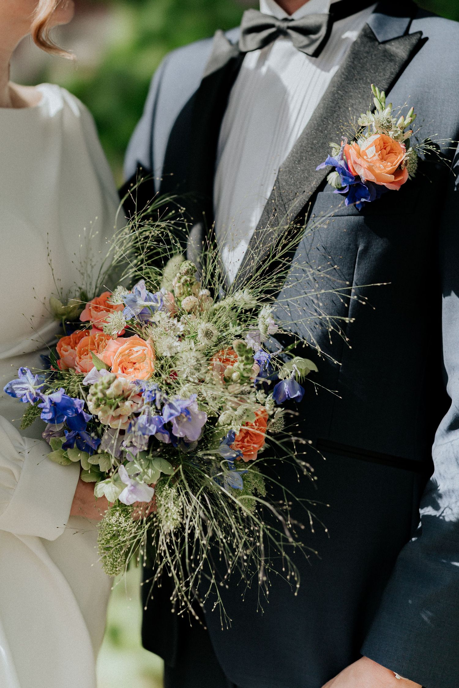 Bride holding a colorful wedding bouquet with orange roses and blue flowers beside a groom in a gray tuxedo with boutonniere.