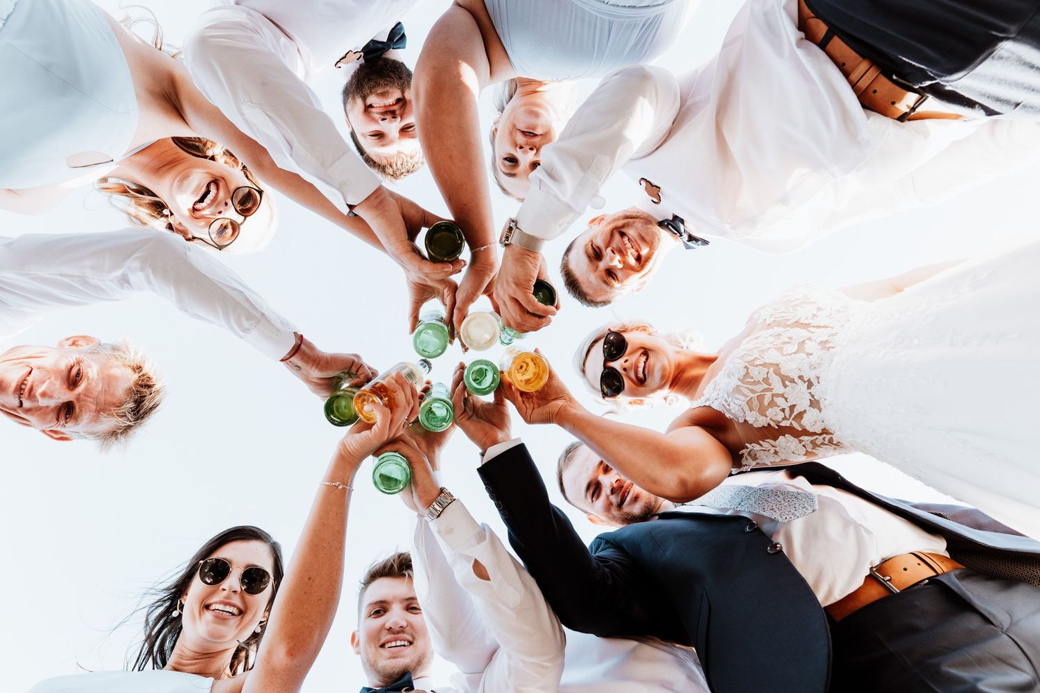 Wedding guests toasting drinks in circle photographed from below against bright blue sky.