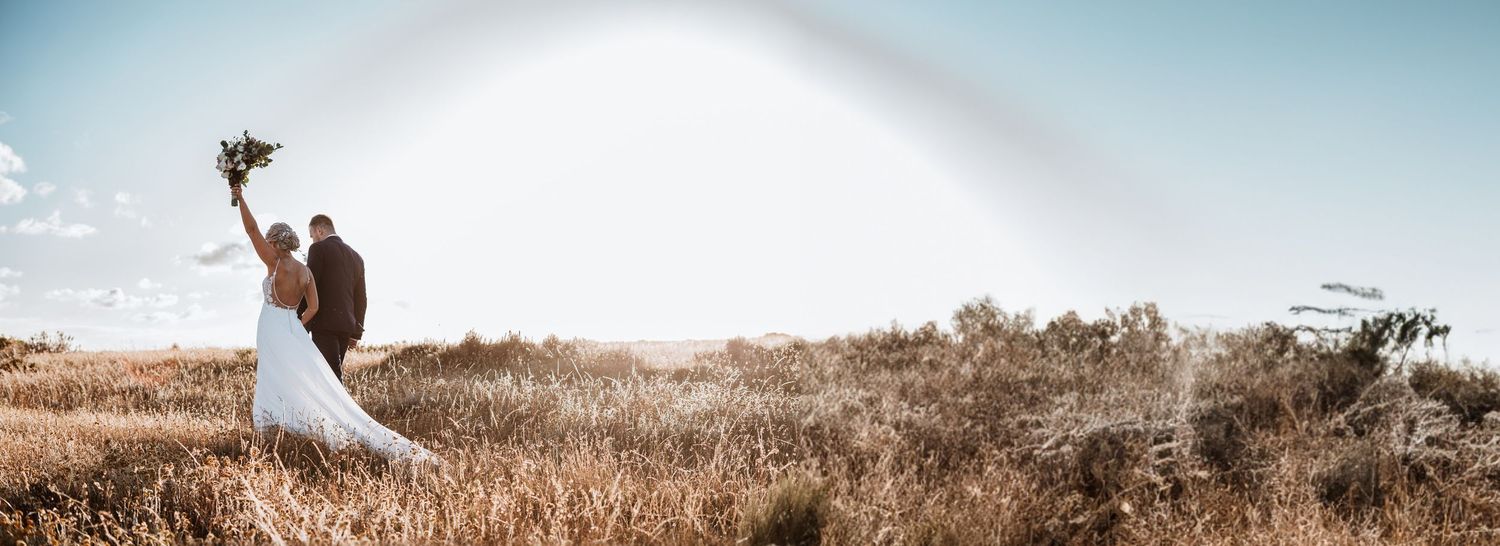 Bride and groom standing in a golden field holding a bouquet under a rainbow.