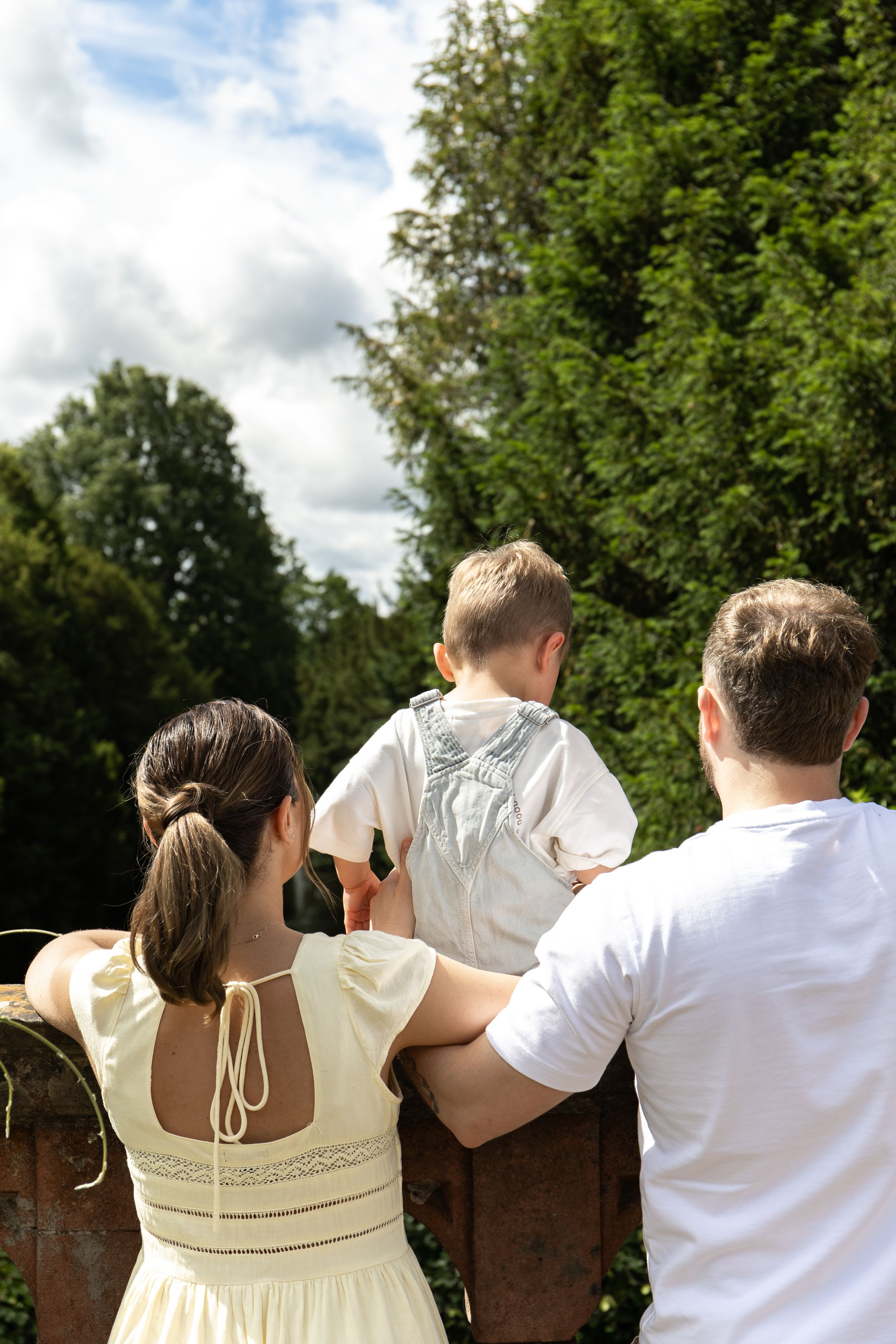 A family of three viewed from behind, looking out at lush green trees on a sunny day natural Outdoor family photoshoot Nottingham