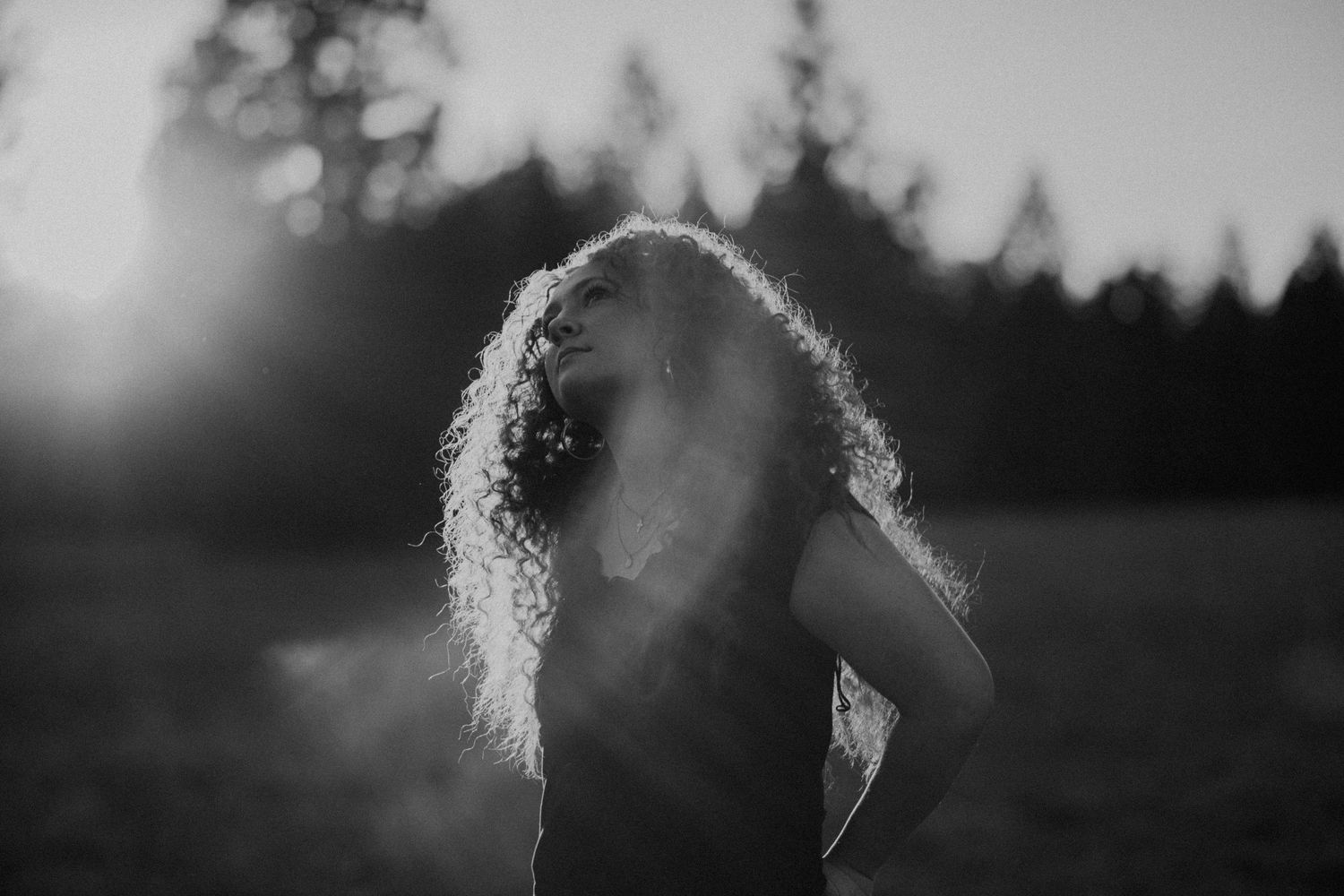 Black and white photo of a woman with curly hair tossing her head back in a sunlit field at dusk.