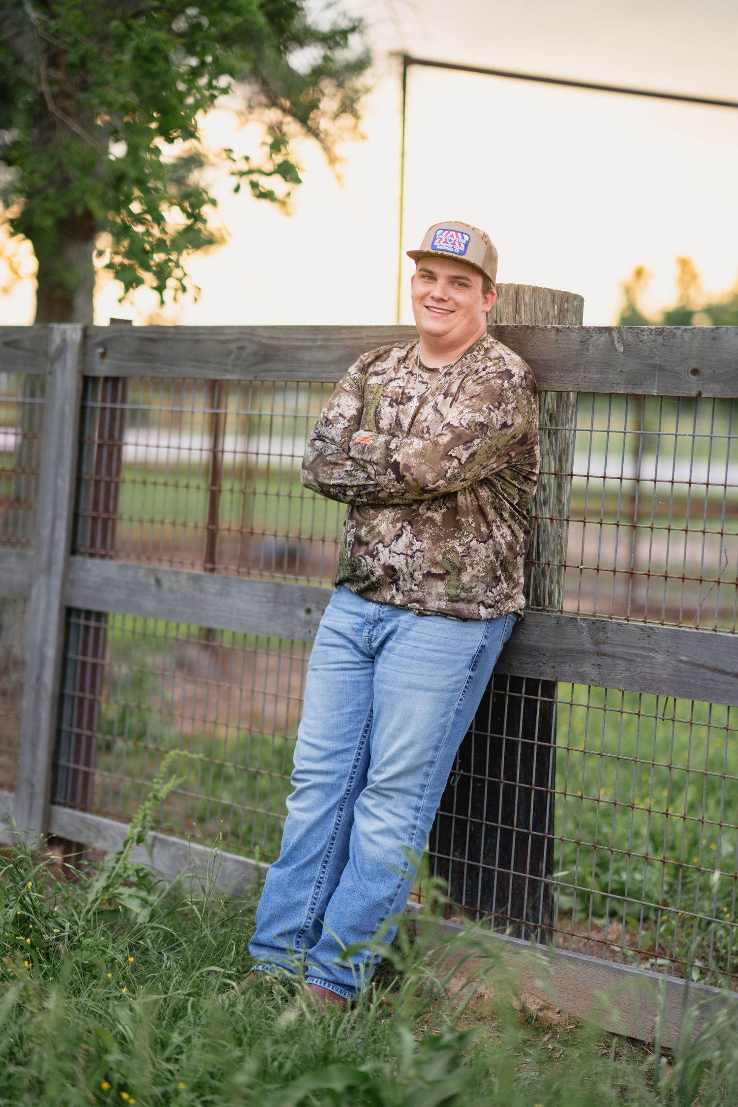 Young man in camo shirt and trucker hat leans against rustic wooden fence at golden hour on a farm.