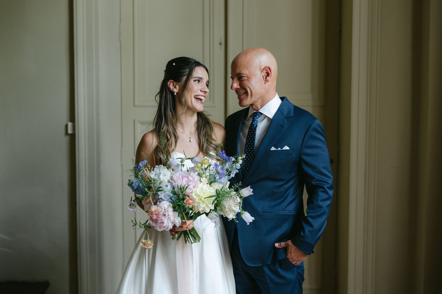 Bride and bridesmaid holding a pastel spring bouquet with pink peonies and blue wildflowers, wearing white and yellow gowns.