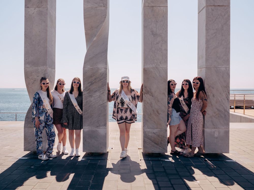 Group of women in bachelorette sashes posing between marble columns by the seaside on a sunny day.