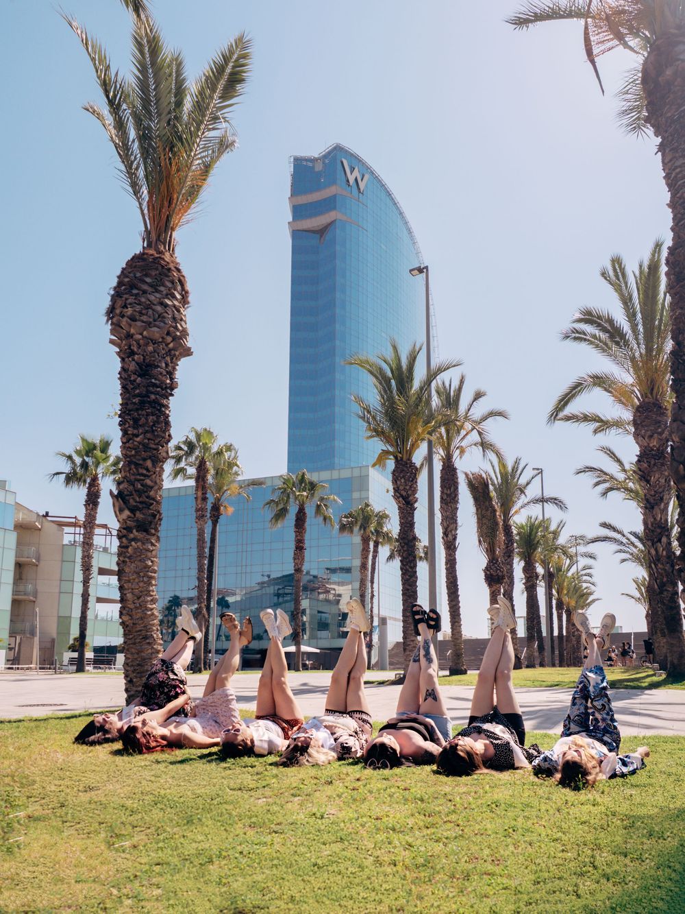 Group of friends lying on grass with legs up near palm trees, W Barcelona hotel skyscraper in background on sunny day.