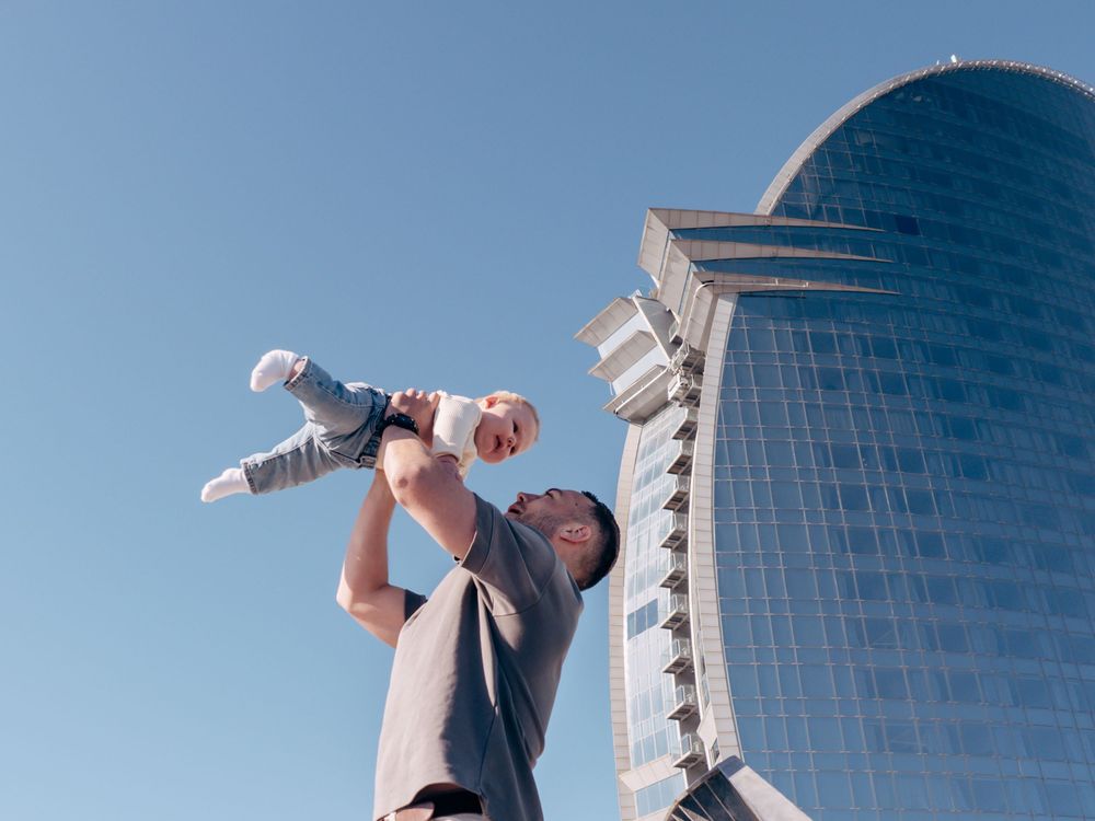 A joyful father lifts his baby high in the air against a clear blue sky near a modern glass skyscraper.
