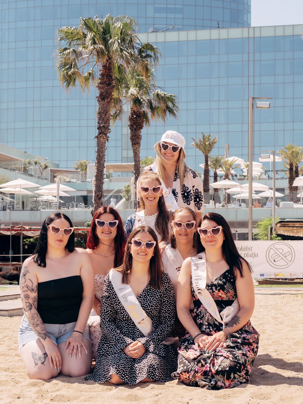 Group of seven women in sunglasses celebrating a bachelorette party on a sunny beach with palm trees and a modern building.