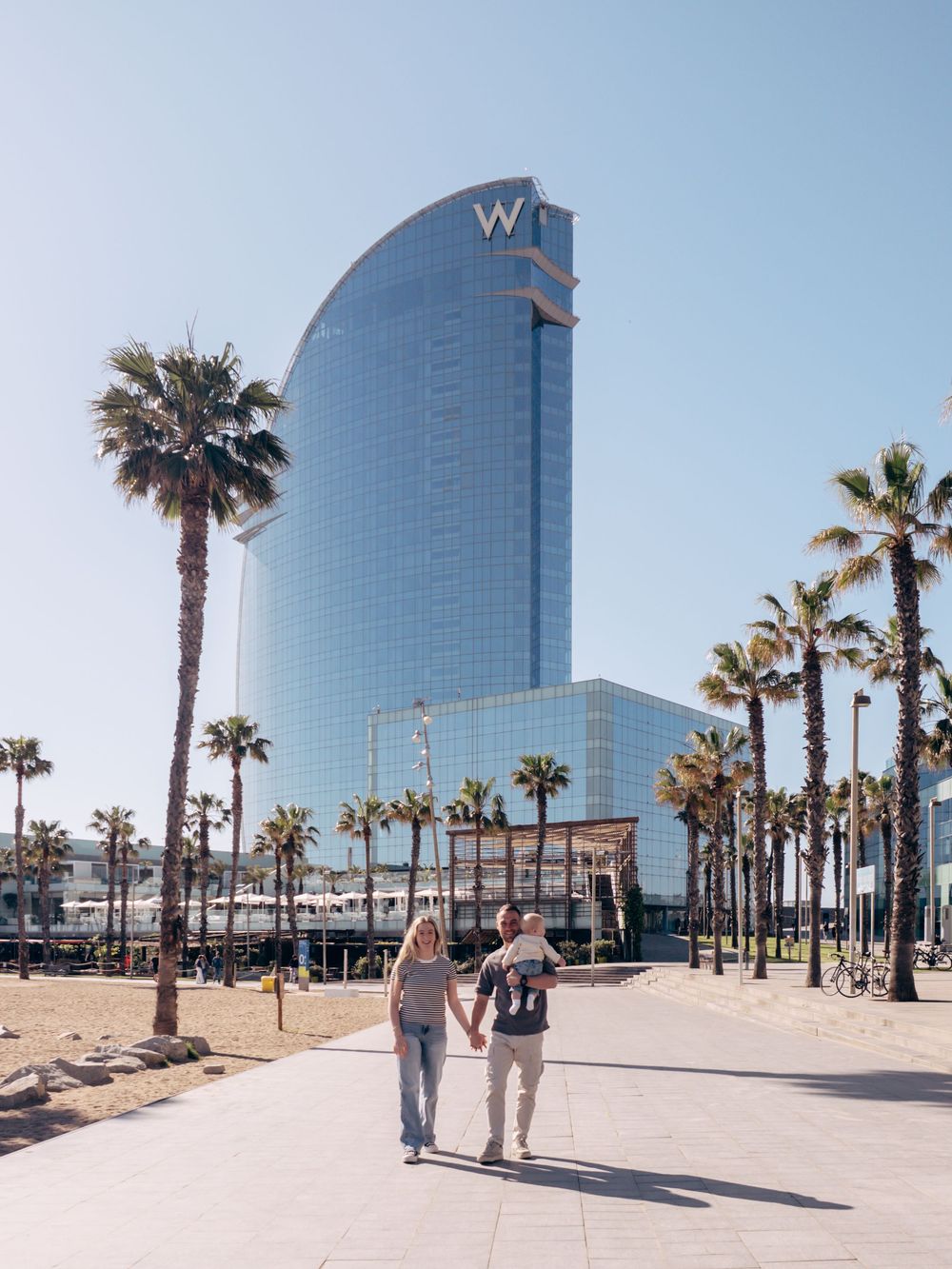 Couple holding hands walking toward the W Barcelona hotel, surrounded by palm trees on a sunny day.
