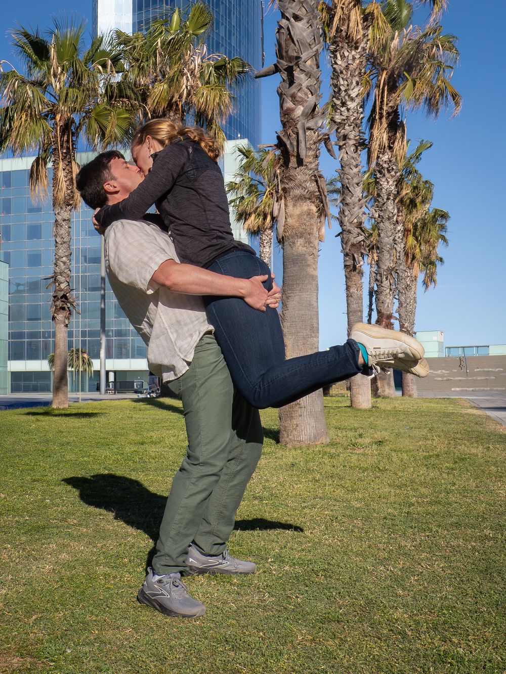 A man lifts a woman in a romantic embrace outdoors near palm trees and a modern glass building on a sunny day.