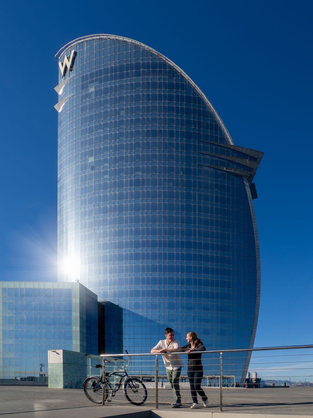 Couple standing by railing with bicycle in front of W Hotel Barcelona's iconic sail-shaped glass skyscraper under blue sky.