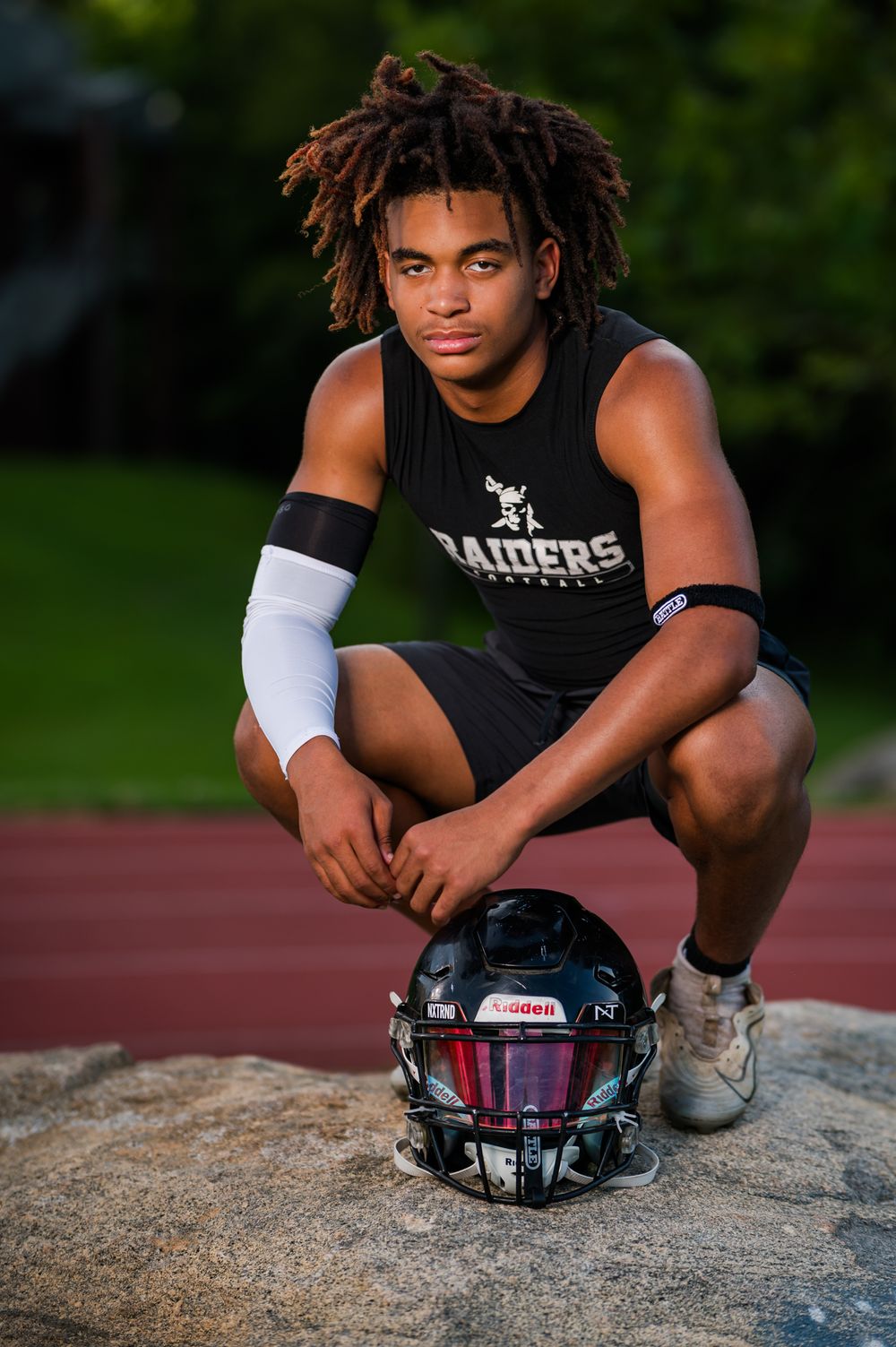 Young football player in Raiders jersey crouching on rock with Riddell helmet, wearing white arm sleeve outdoors.