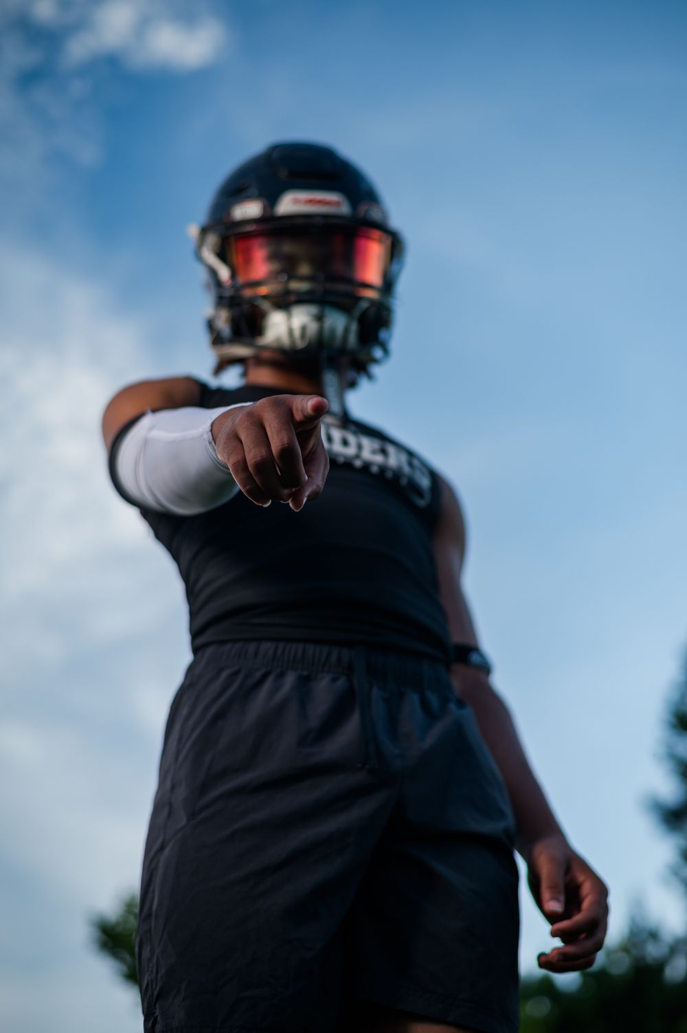 A football player in black uniform and helmet points toward camera against a blue sky background.