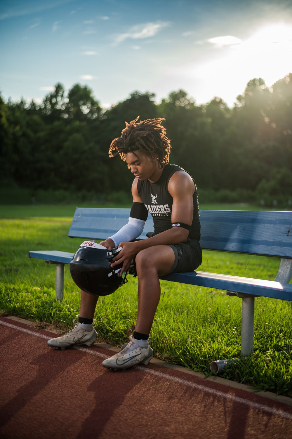 A young football player sits on a bench holding his helmet, wearing a Raiders jersey and cleats, with a sunset backdrop.