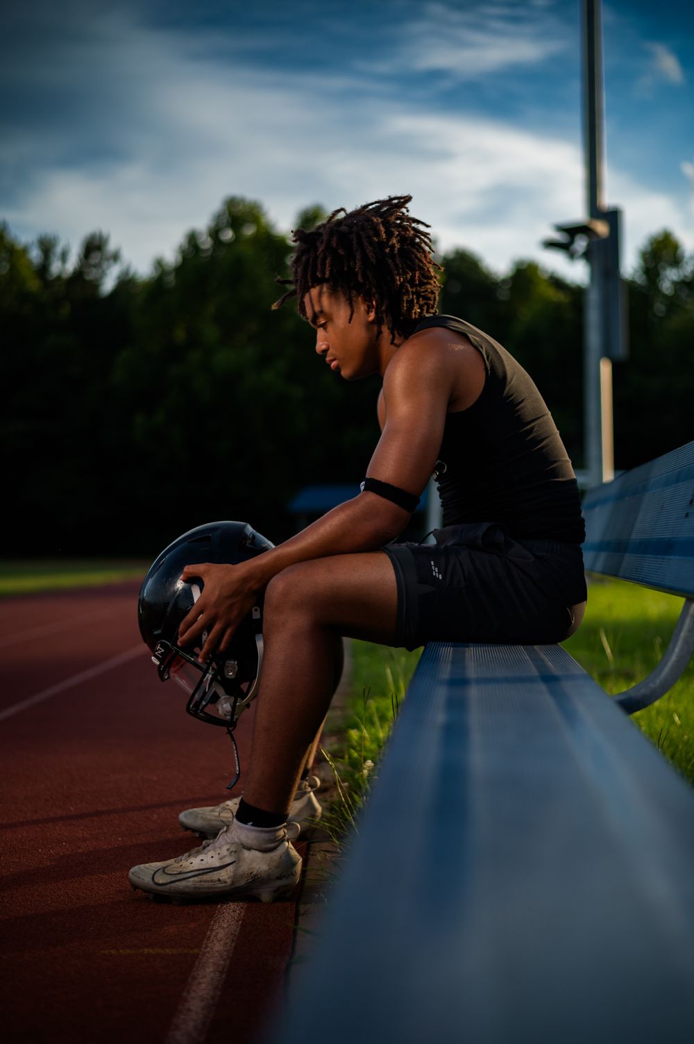 Young football player sitting on bench holding helmet, wearing cleats and tank top on outdoor track at dusk.