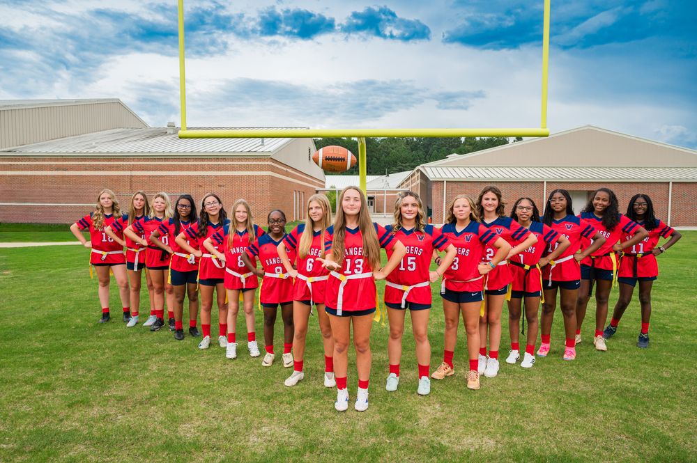 Girls flag football team poses on green field in red uniforms with goalposts and brick building backdrop.