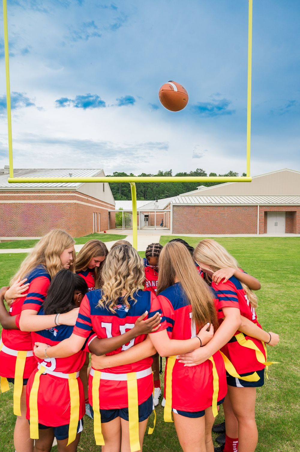 Girls flag football team huddles together on field with football in air near goal post.