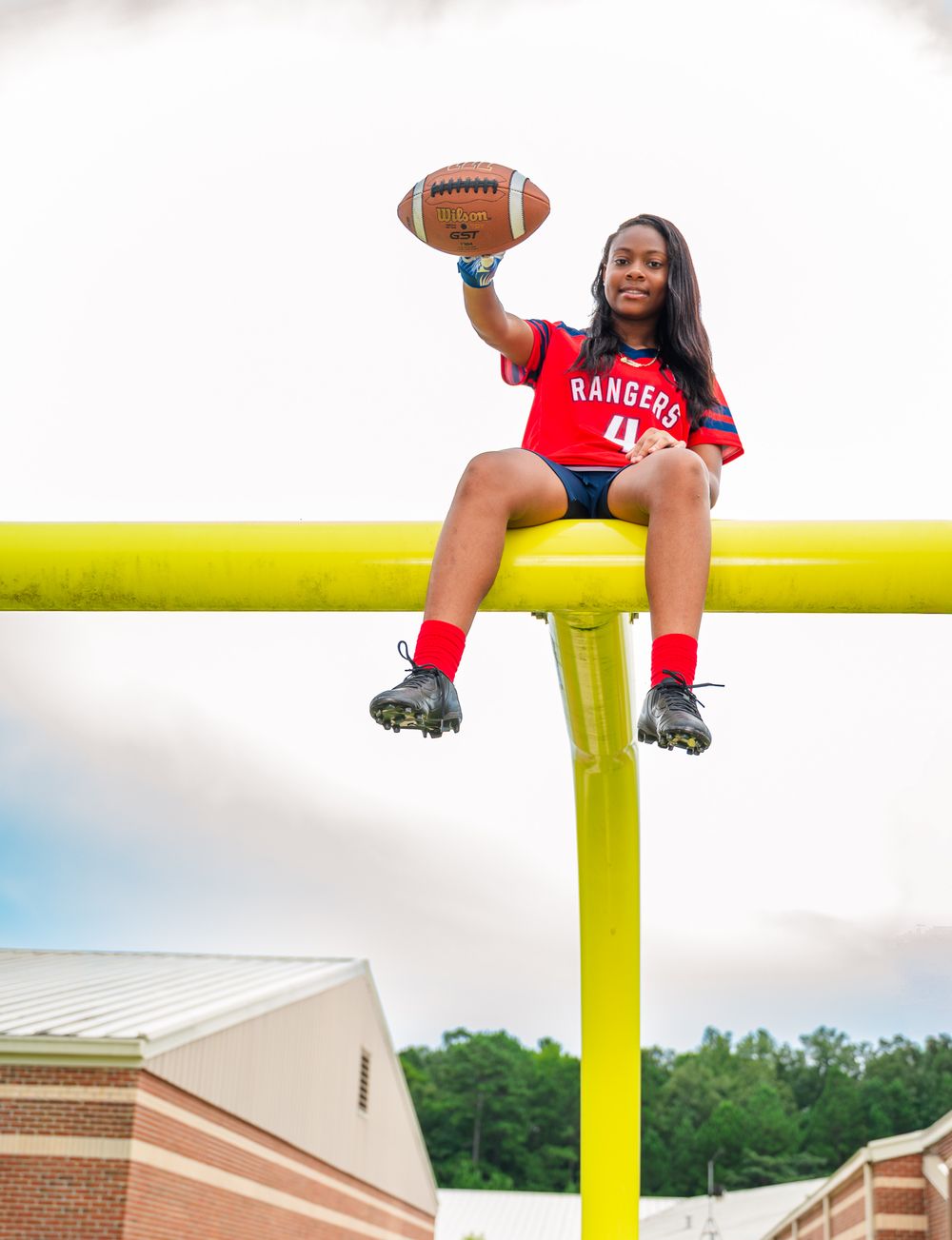 Young female football player in Rangers jersey sits atop yellow goal post holding football aloft outdoors.