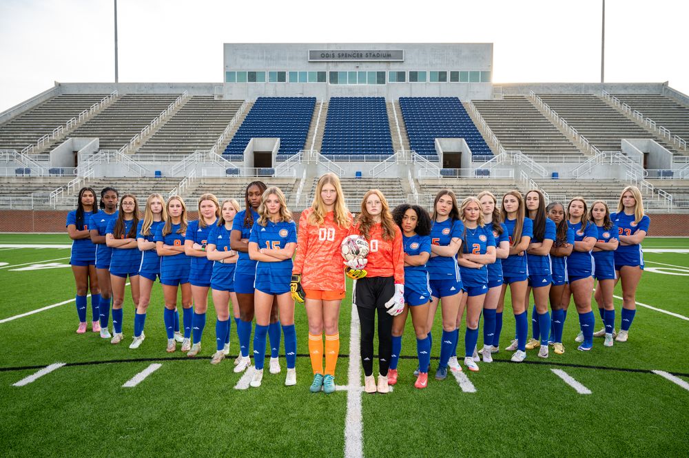 Girls soccer team poses in blue uniforms on stadium field, with goalkeeper in orange at center.