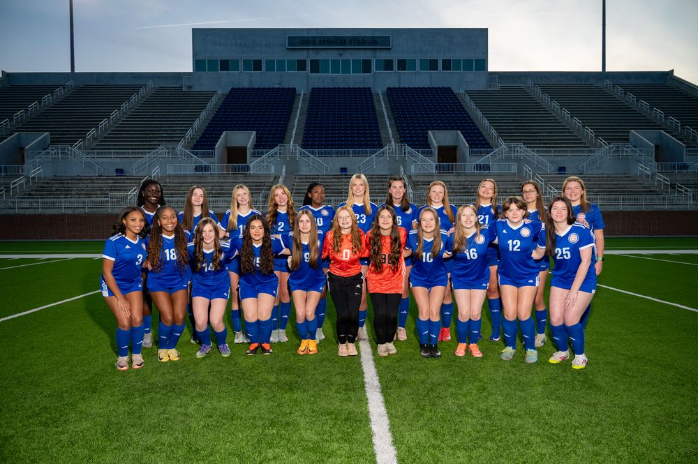 Girls soccer team poses in blue uniforms on a stadium field with bleachers in the background.