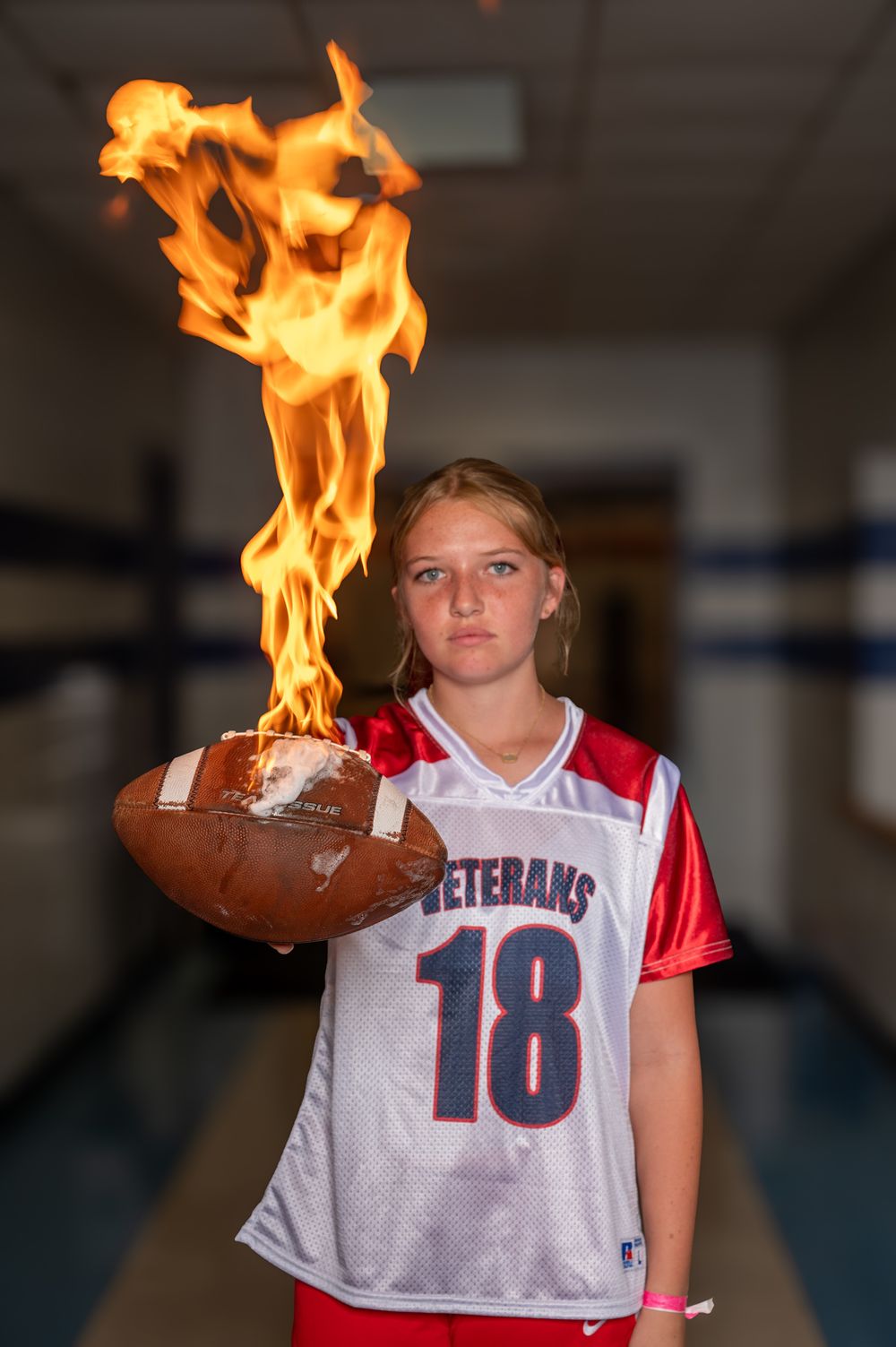 Girl in Veterans #18 jersey holds a flaming football in a school hallway.
