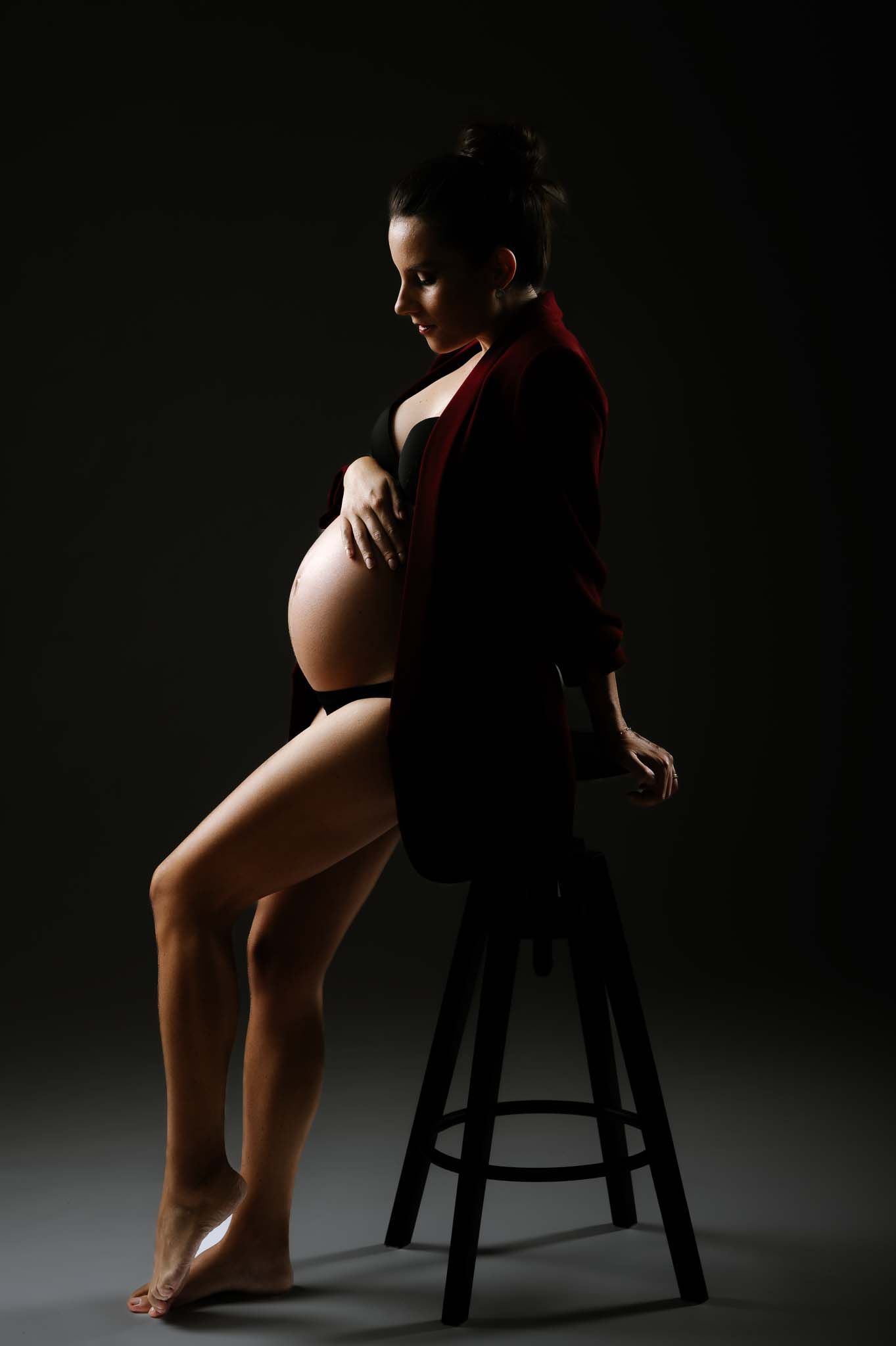 Pregnant woman in black outfit sitting on stool, cradling baby bump in dramatic studio lighting.