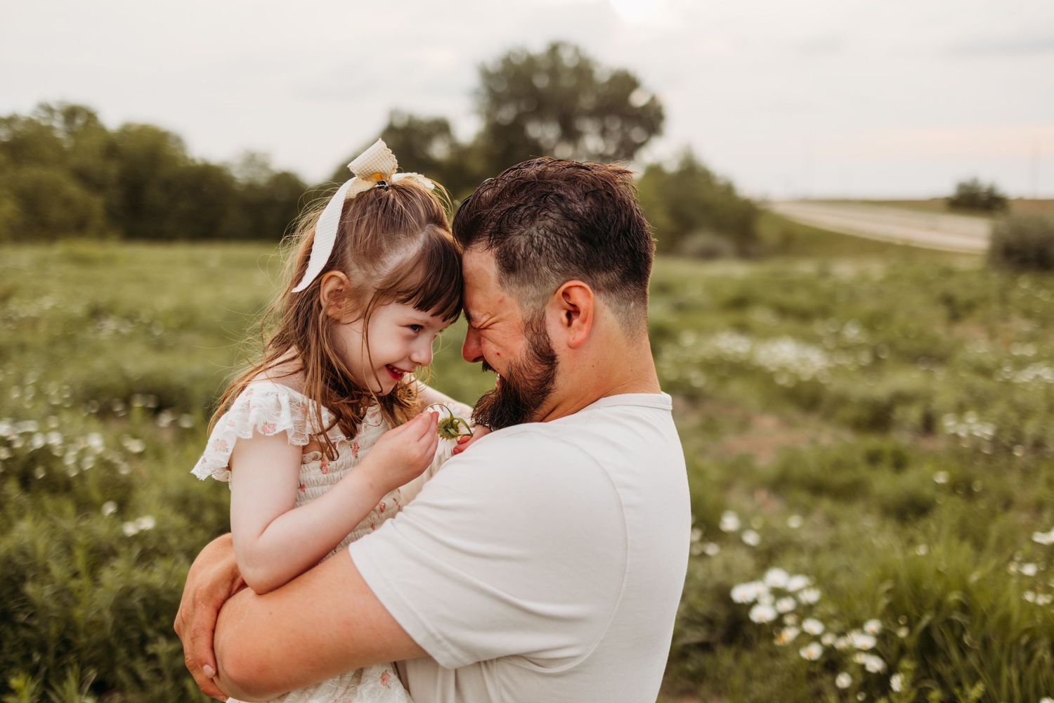 A father holds his young daughter in a flower field, their foreheads touching as she holds a small wildflower.