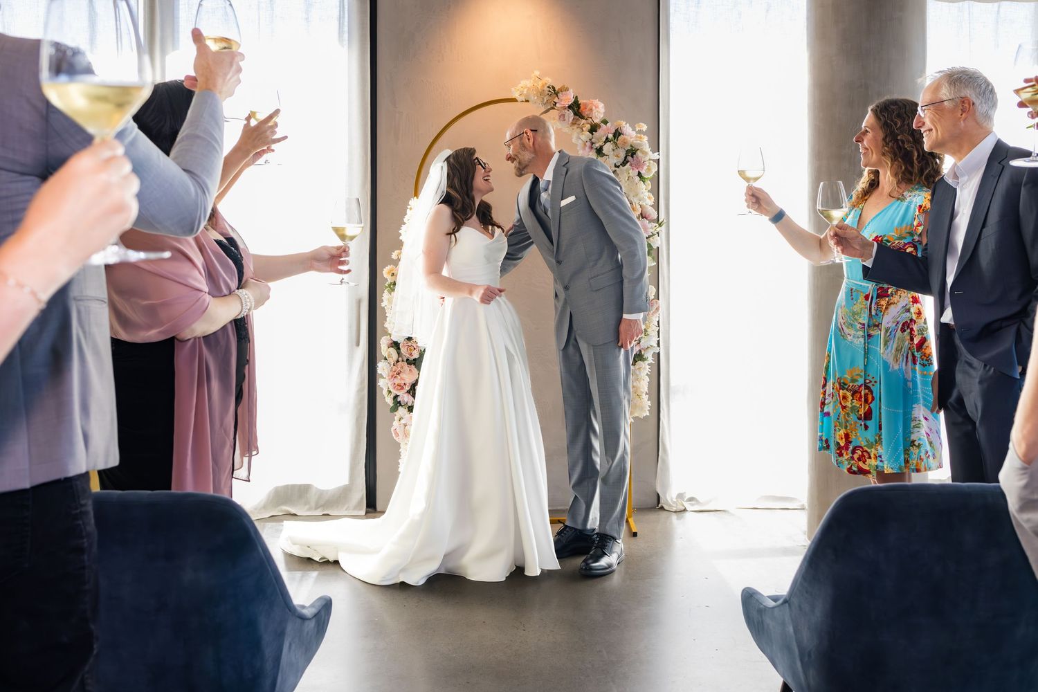Bride and groom share first kiss at indoor wedding ceremony while guests raise glasses in a toast around them.