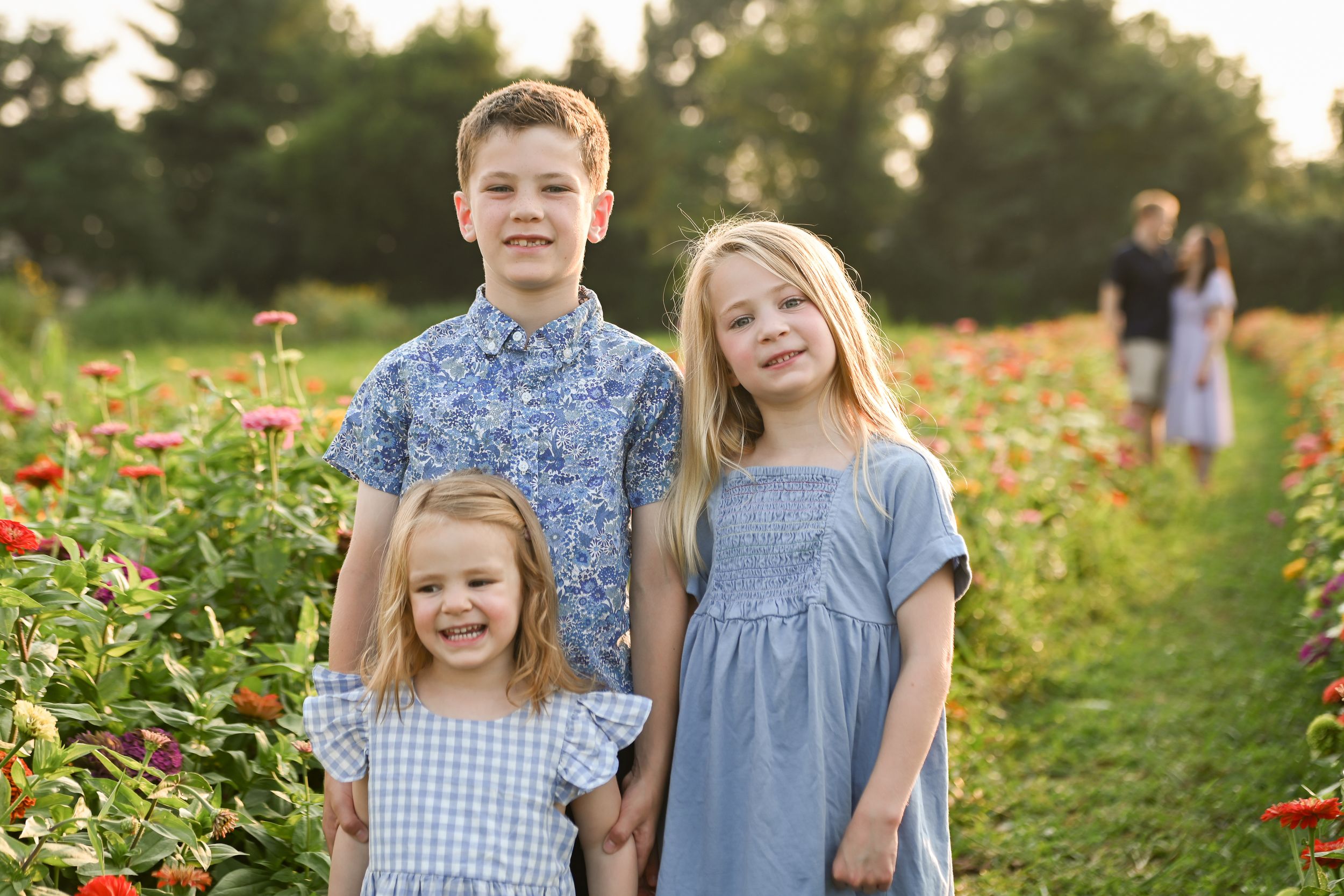 Three children smiling in a colorful flower field, two girls in blue dresses and a boy in a floral shirt.