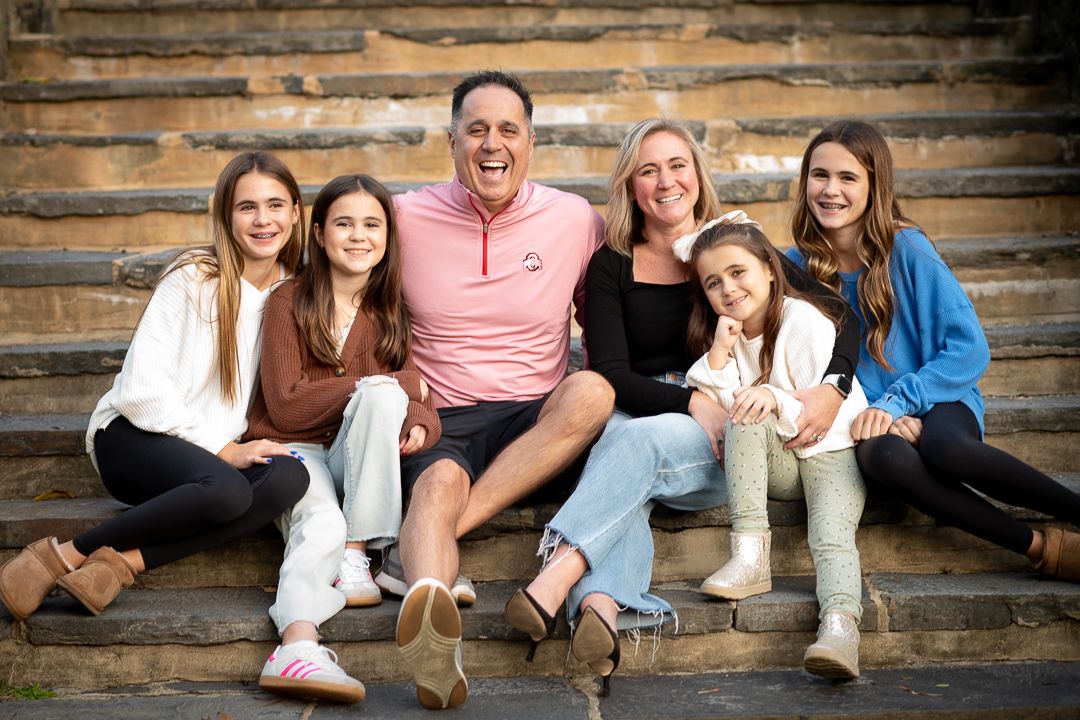 Happy family of six smiling together on stone steps, featuring parents and four daughters in casual attire.