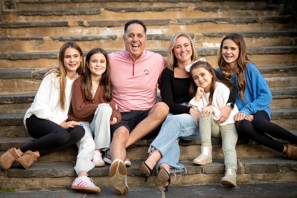 Happy family of six smiling together on stone steps, featuring parents and four daughters in casual attire.