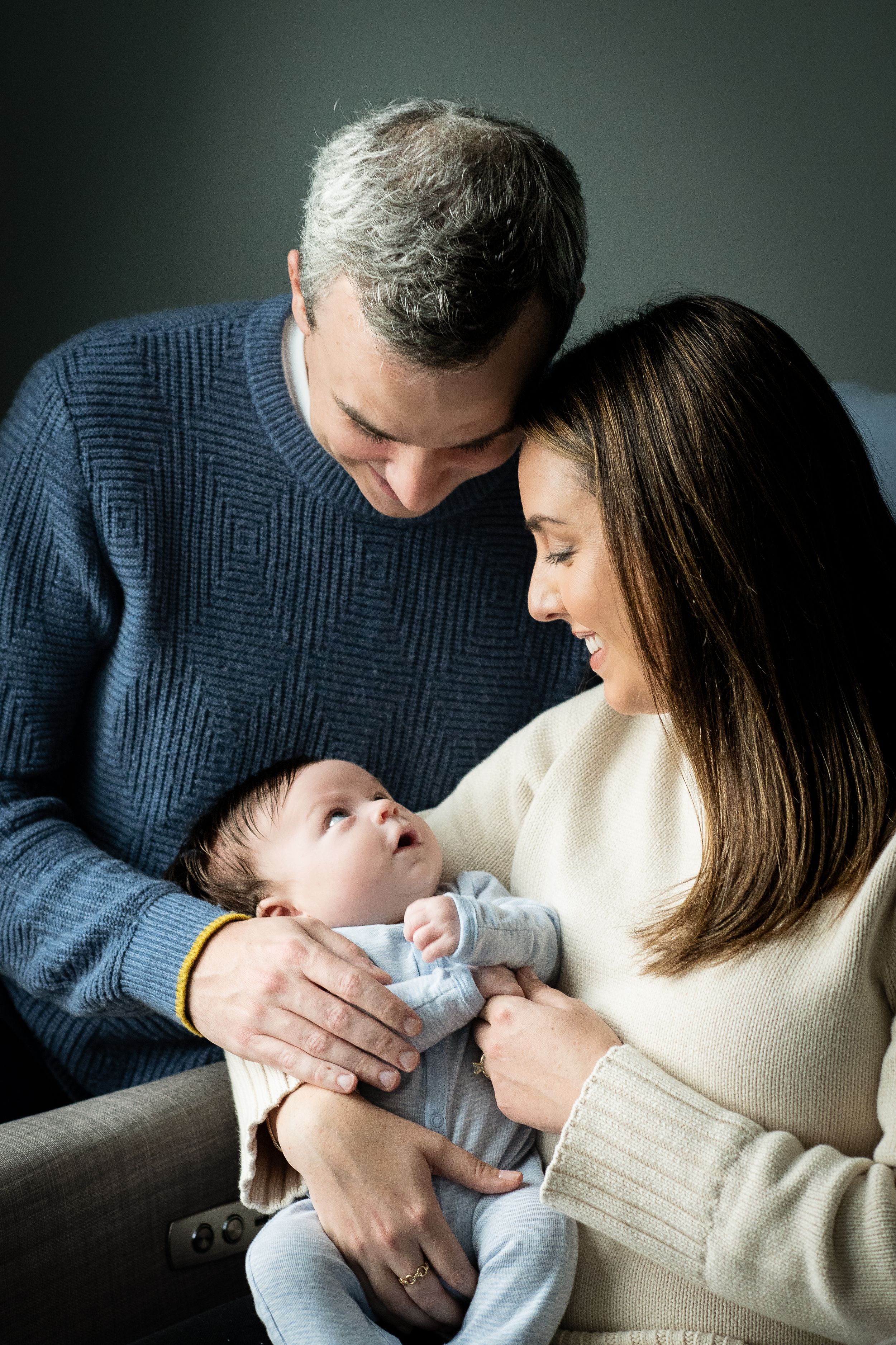 A couple smiles lovingly at their newborn baby, with the man in a blue sweater and woman in cream top.