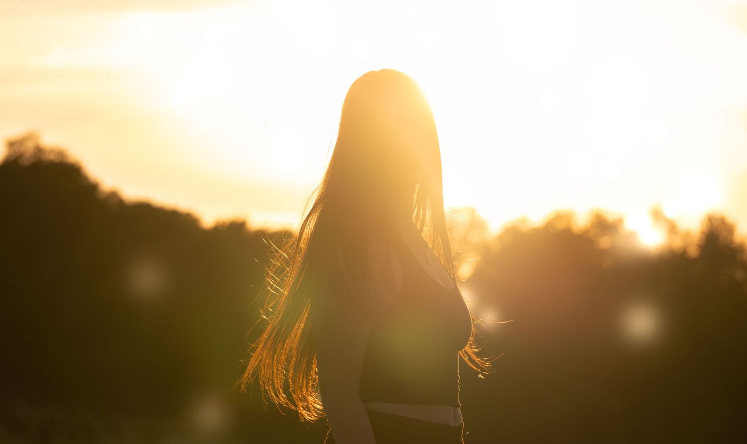 Silhouette of a woman with long hair standing in a golden sunset with warm sunlight and bokeh effects.