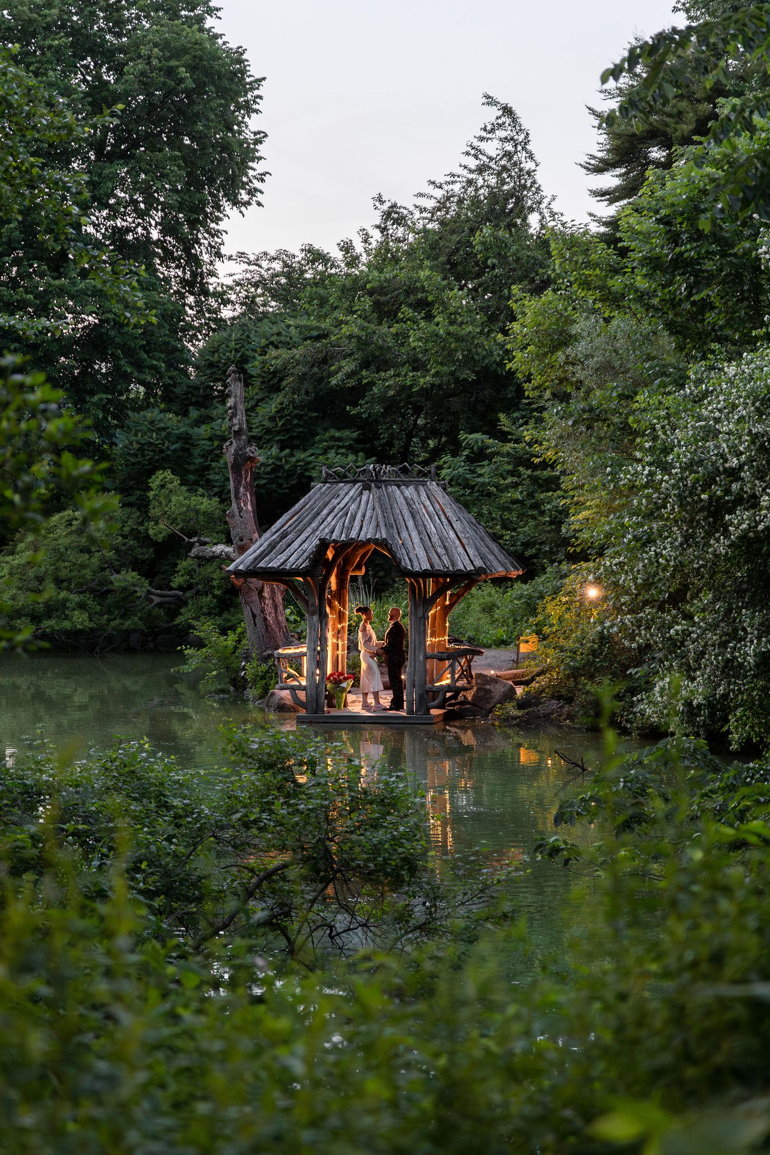A romantic wooden gazebo glowing warmly at dusk, nestled among lush green trees beside a tranquil reflective pond.